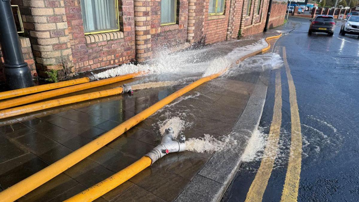 Water pumping out onto a path and road from five yellow hoses. There are red brick houses along the left of the path. 