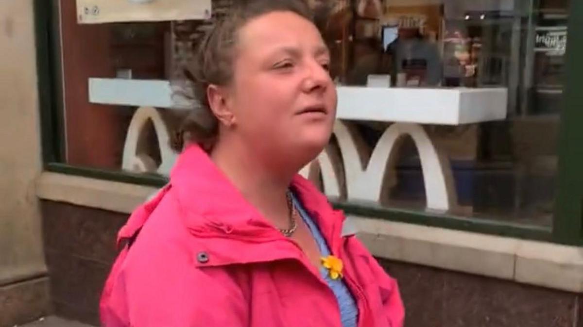 A young woman in a pink coat and brown hair tied back looks off camera.