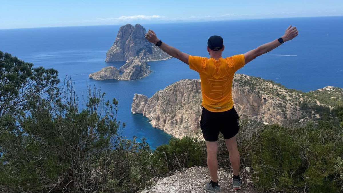 Mike Jones with his back to the camera, standing on an edge with his arms out looking at the sea and view in Ibiza in April 2025.