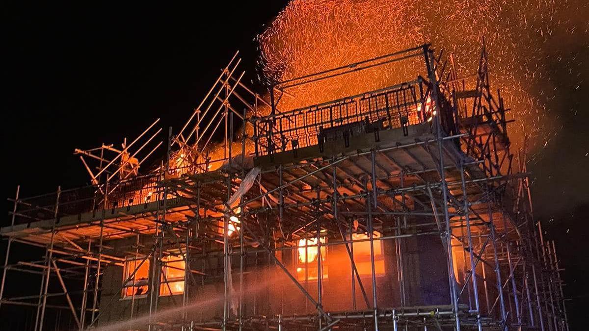 A house under construction surrounded by scaffolding is well alight and red flames and smoke and embers can be seen enveloping it and glowing against a night sky.