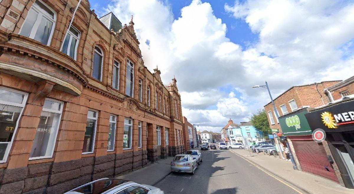 An image taken from the road of Boston Borough Council building.  It is a red stone building with handsome white windows and ornate finishing touches on the roof.  Opposite is a shop with it's grill down and a car park.