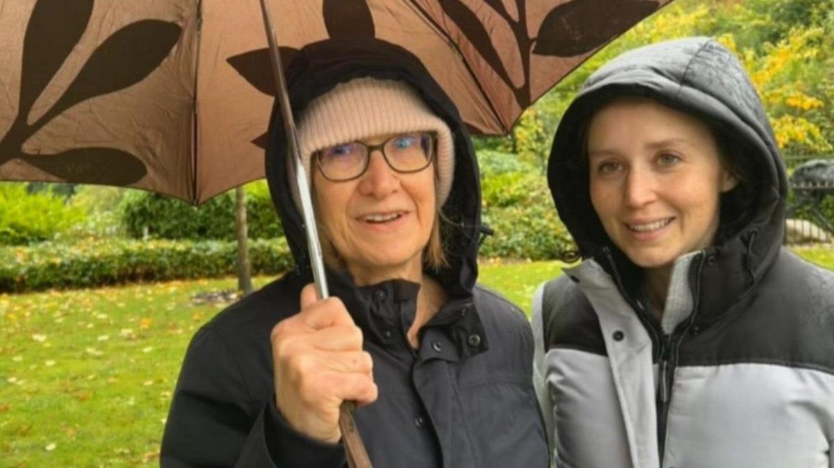 Two women, one on the left wearing glasses and holding an umbrella, with one on the in a black and grey coat