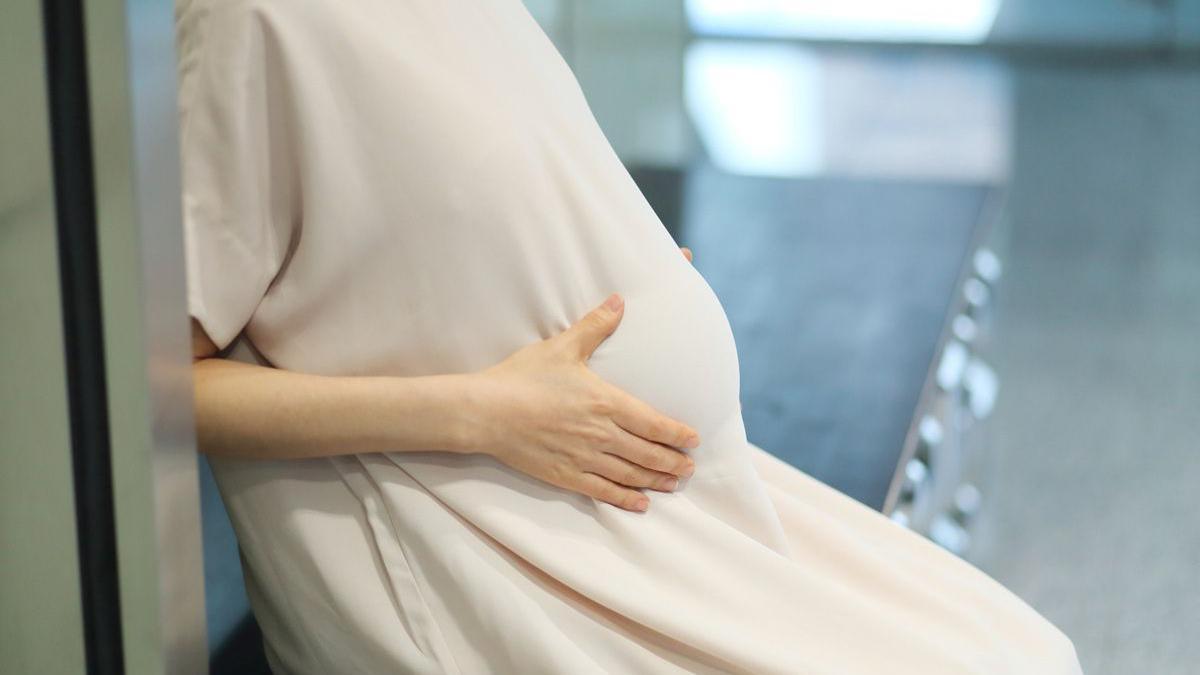 Close-up of a pregnant woman's torso, with her hands cupping her baby bump. She is wearing a long beige dress and sitting on a bench.