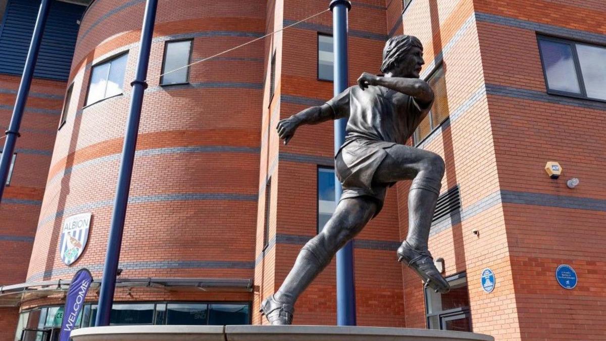 A bronze sculpture depicting a man wearing football kit, including football boots, with one foot totally and the other partially off the ground, with studs on the boots showing. The outside of the stadium is visible behind the statue, with the West Bromwich Albion crest on the bricks.
