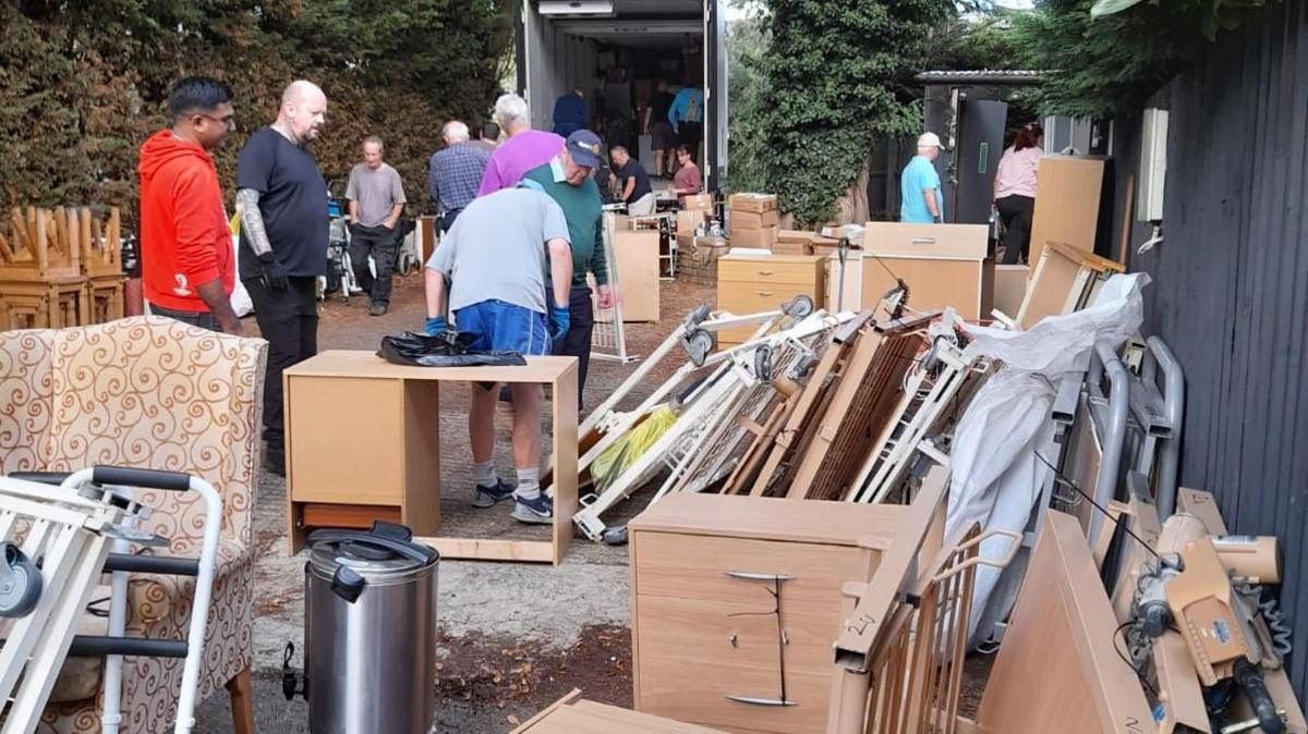 Various items of furniture including a brown armchair, a wooden desk with drawers, a tea urn, and several bed frames. There are people amongst the items and a lorry with its back opened is visible in the background.