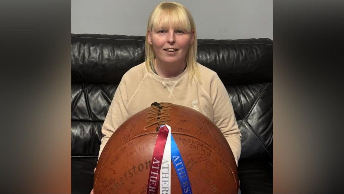 A woman with shoulder-length blonde hair sits on a black sofa, wearing a beige top. She holds a brown ball on her lap with stitches along the top holding the leather together. Three ribbons are attached to the top of the ball, blue, mauve and white, each with the word "Atherstone" on them.