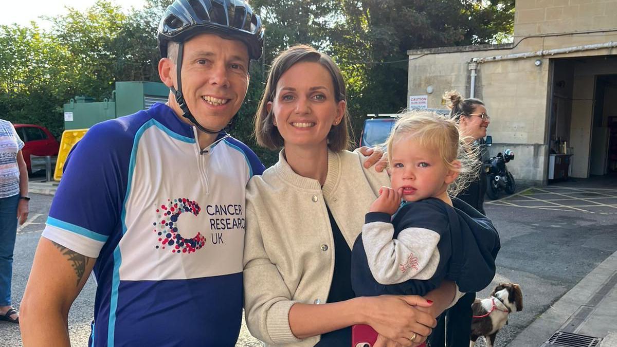 Mark Parsons stands next to his wife, Jess Parsons, who is holding their baby daughter