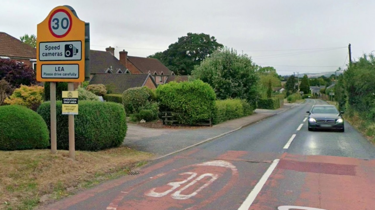 A yellow road sign with a 30mph limit and the words speed cameras, and Lea please drive carefully on it. The sign is to the left side of the road, with houses and hedgerows to the left, and a car approaching the camera on the right.