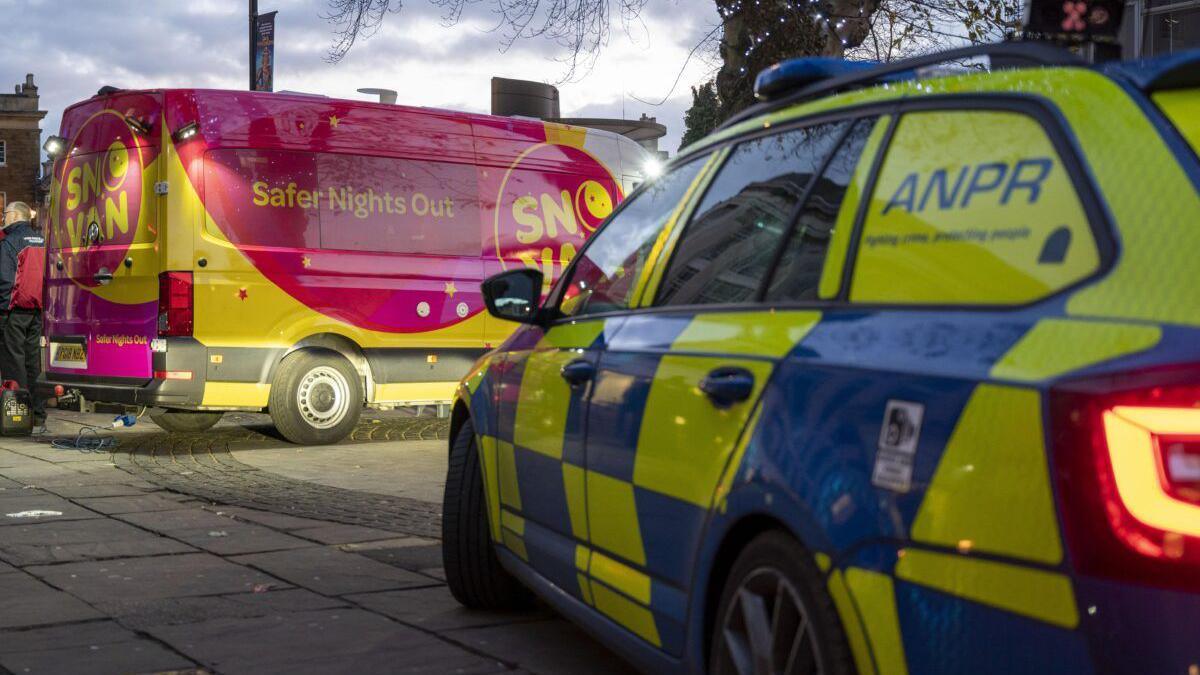 A Safer Nights Out bus, by an ANPR vehicle, in the middle of a town.