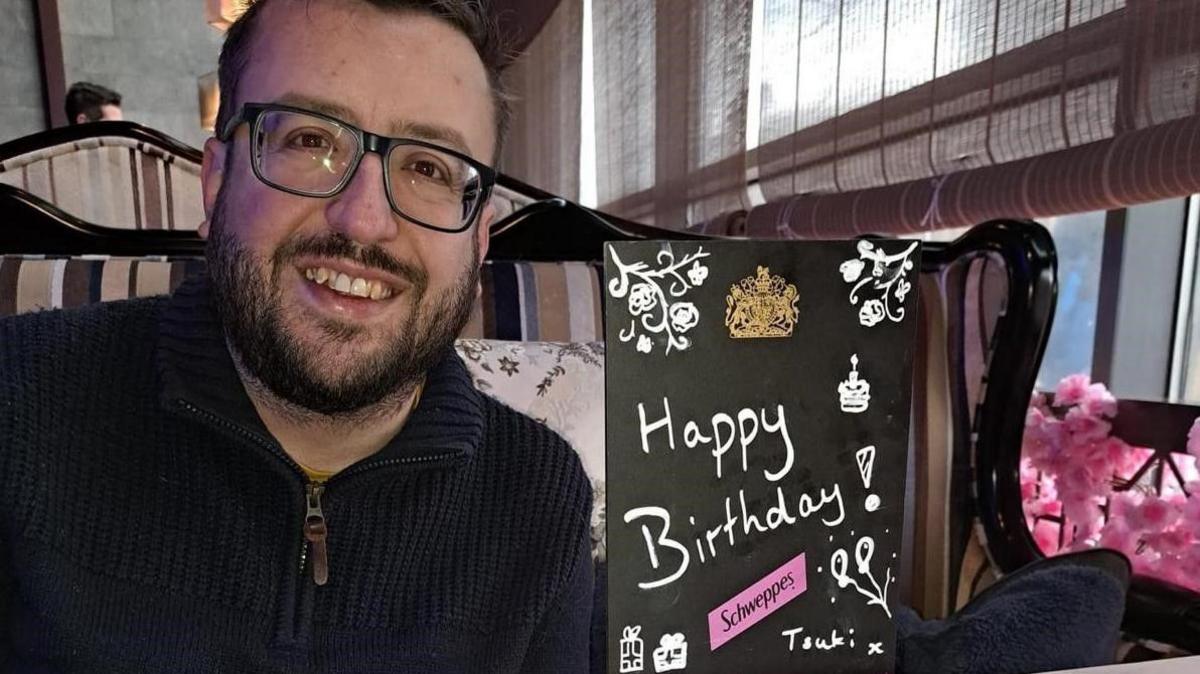 A white male with dark brown hair and beard wearing a navy zip up top sits next to a black sign saying 'Happy Birthday'