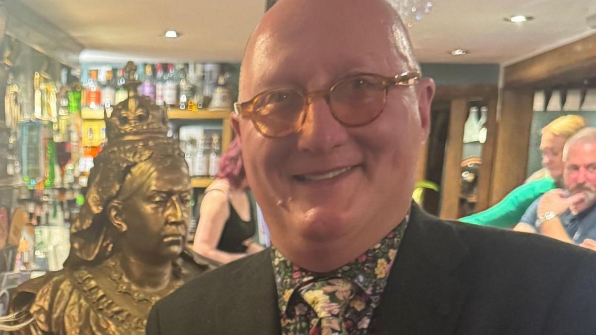 James Burrell is wearing a floral shirt and a florla tie with a black blazer. Behind him in the pub is the gold bust of Queen Vistoria.