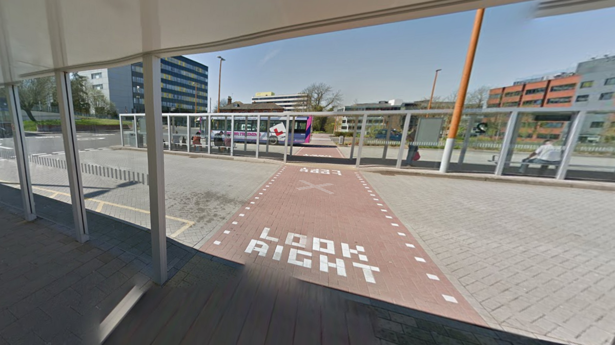 A bus station comprised of several glass bus shelters, with a red brick pathway which reads "look right".