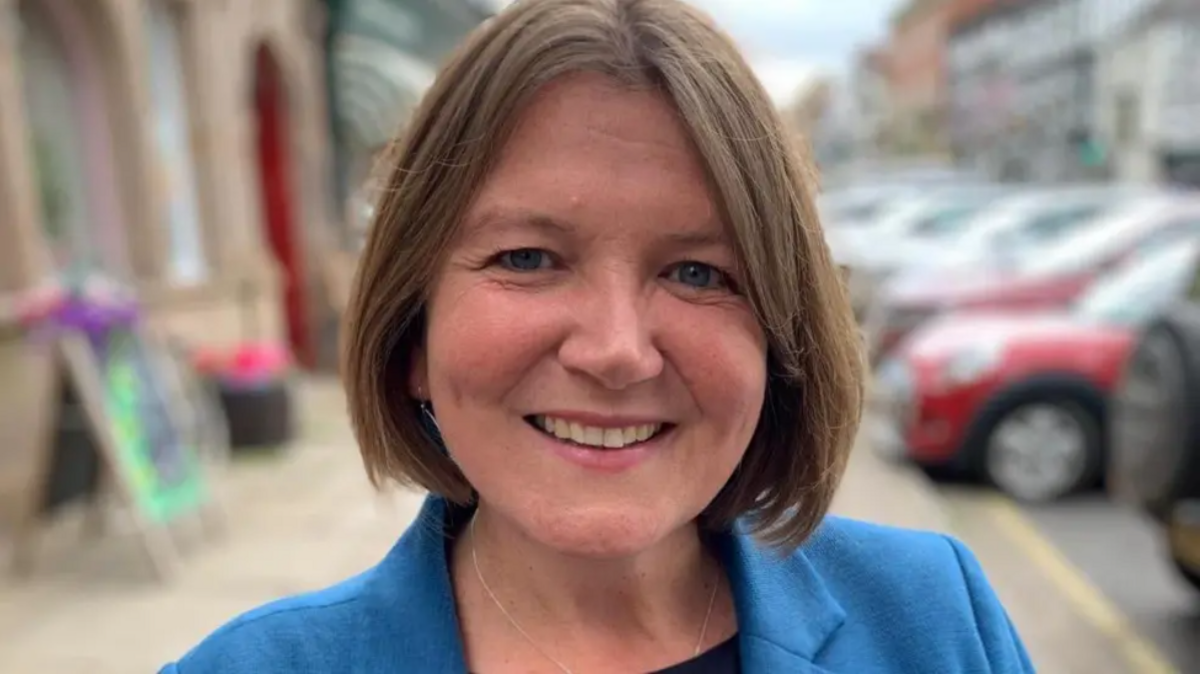 MP Ellie Chowns with a brownish bob haircut wearing a blue blazer and smiling. There are cars blurred in the background. 