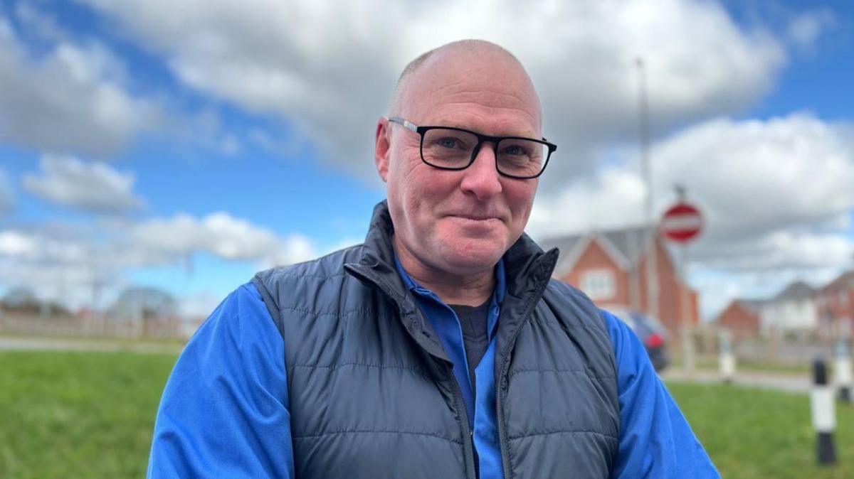 A man with a bald head, blue top and dark blue gilet standing outside on a grassy area