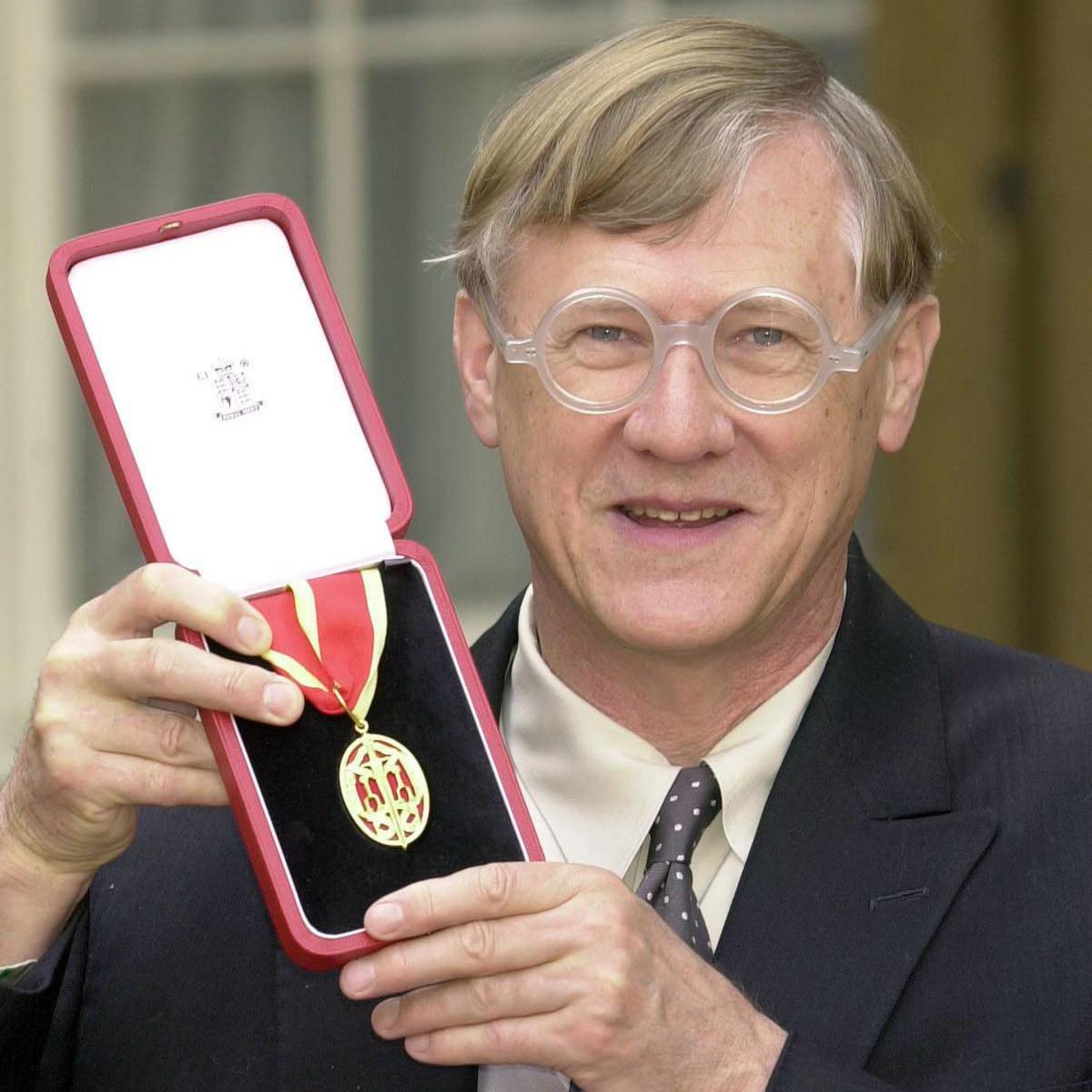Sir Nicholas Grimshaw is holding an open box with a medal on a ribbon to the camera as he smiles. The medal is a gold oval shape with red patterning that forms a crown. The ribbon is red and gold striped. Sir Nicholas is wearing a smart black jacket, a pale grey waistcoat, a cream shirt and a black and cream spotted tie. He has straight blond and grey hair and is wearing a pair of glasses. Behind him are windows.