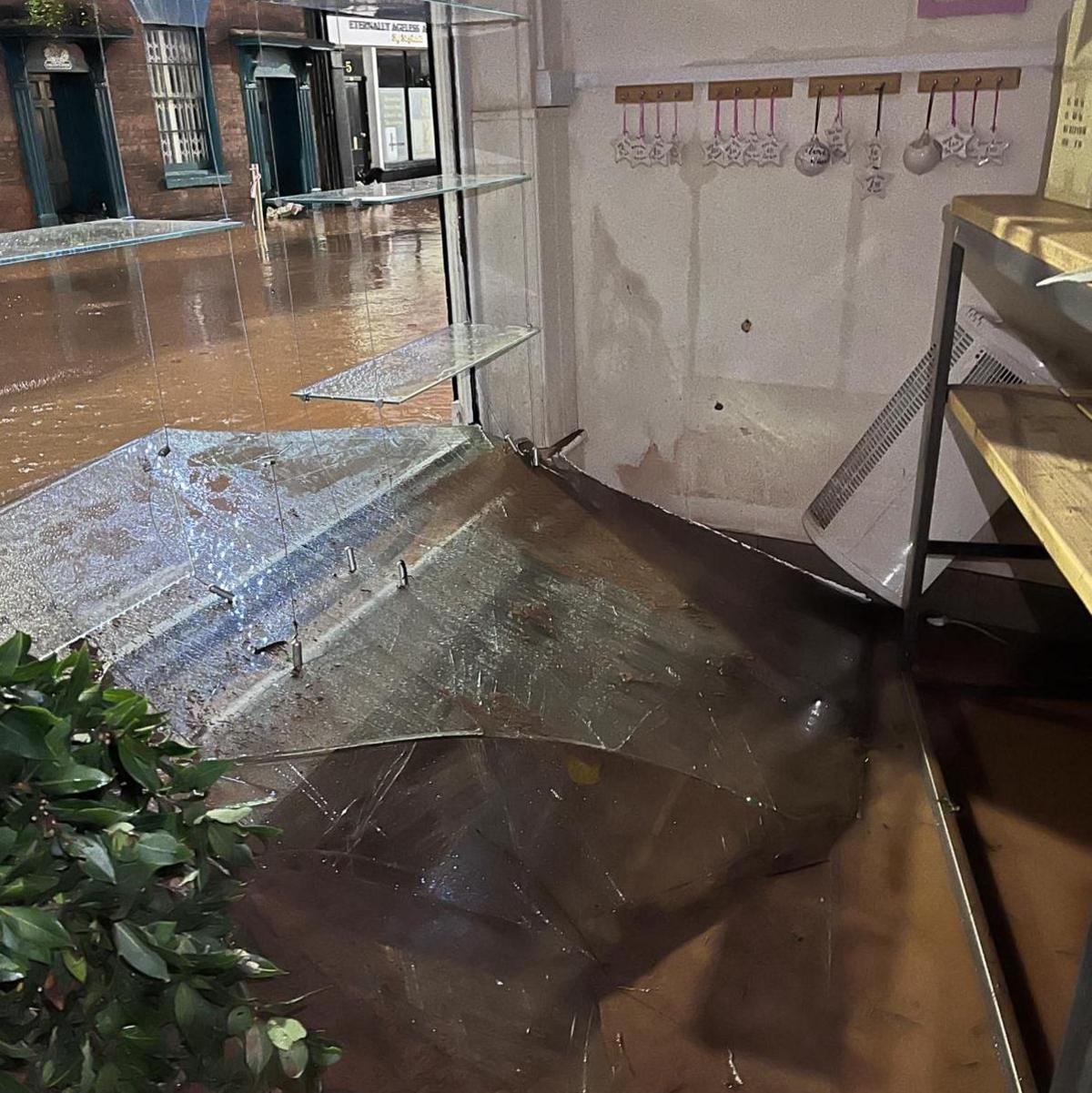 The front window of a shop which has broken and fallen inside the store on to the floor. Flood water is on the floor and can be seen outside on the high street.