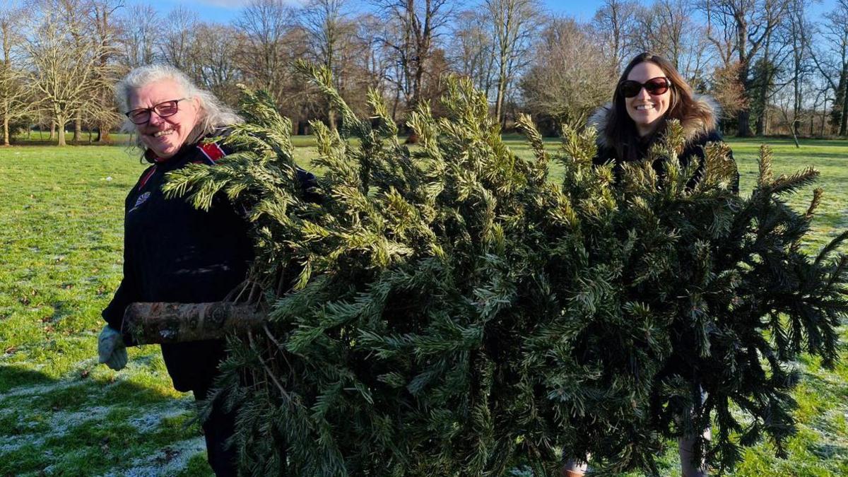 Two volunteers - one on the left wearing glasses and a black jacket with a red strip- holding a Christmas tree with another lady standing on the other side- holding the top of the tree 