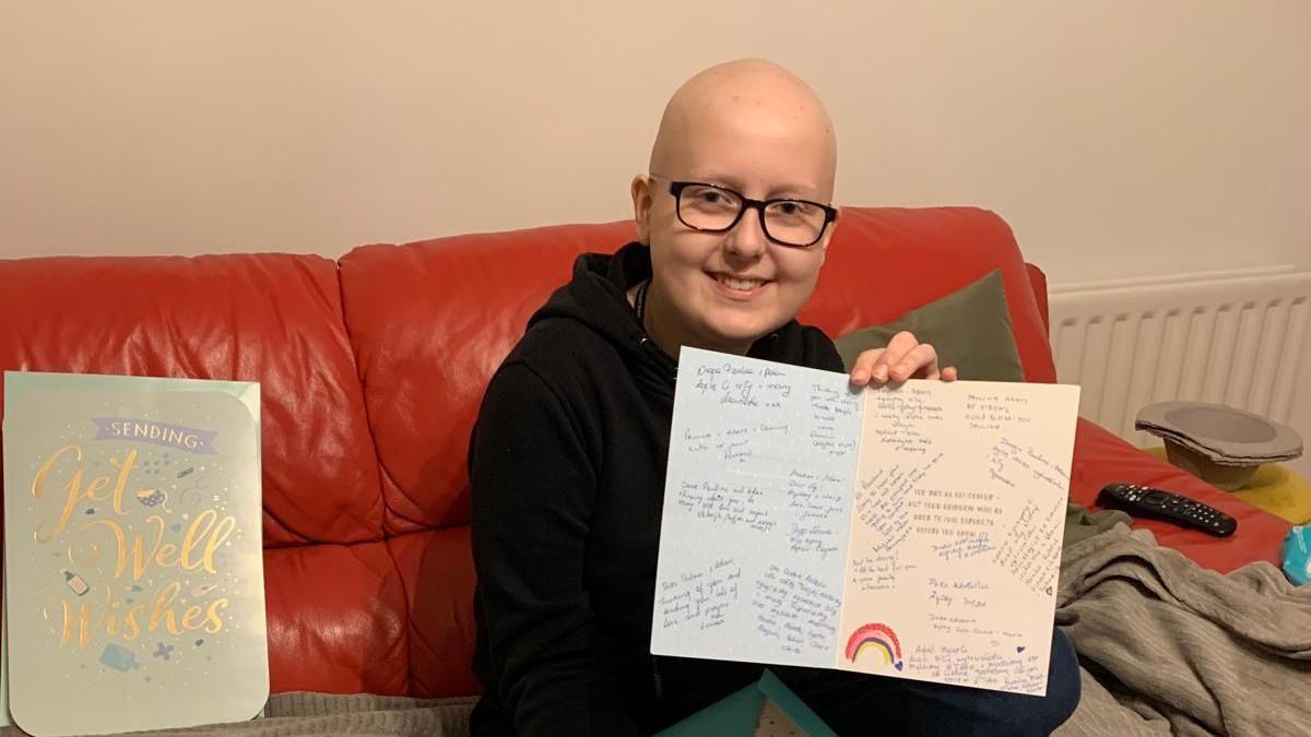 Adam is sitting on a red sofa holding 'get well soon' cards and smiling at the camera. He is wearing glasses and has a shaved head.