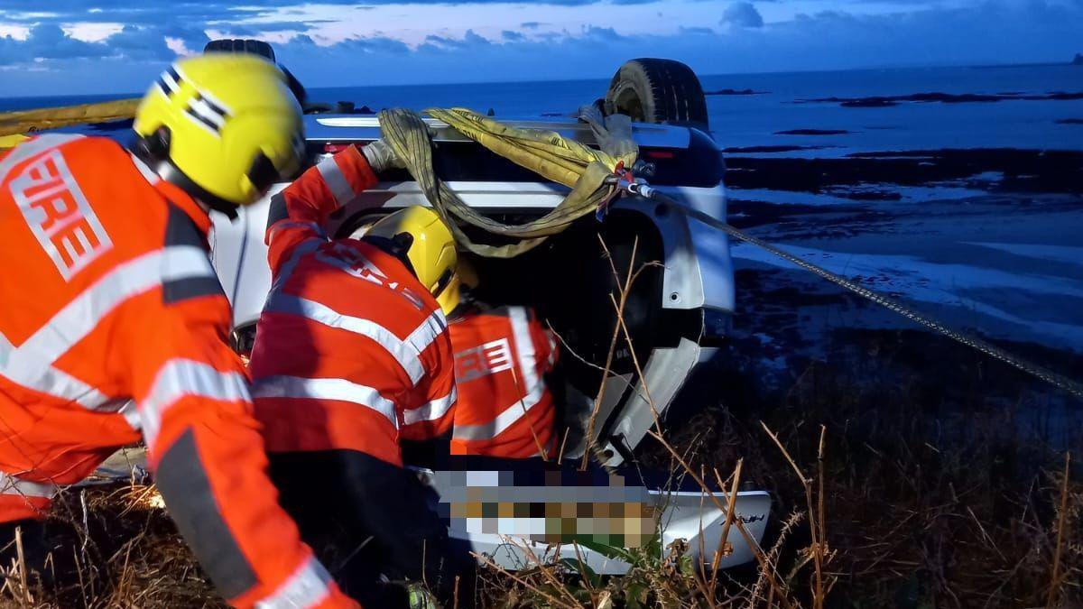 Three fire service crew members attending to the overturned vehicle at La Pulente. The crews are wearing orange hi-vis jackets and yellow hats. 