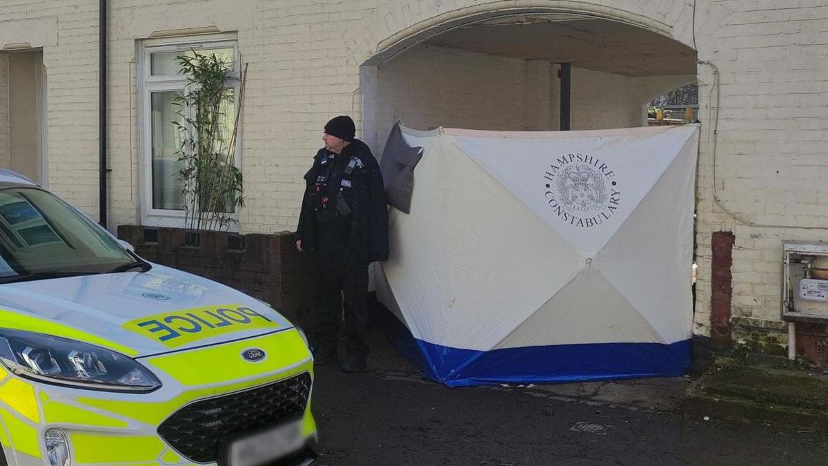 A police officer stands outside an archway between terraced homes in Union Road. A large white screen blocks the archway.