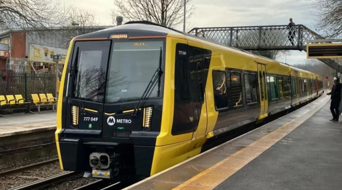 A Merseyrail black and yellow electric train on the track. A bridge over the platform can be seen above the train with a man walking across the other side. One man can be seen standing on the train platform at the bottom looking towards the train.