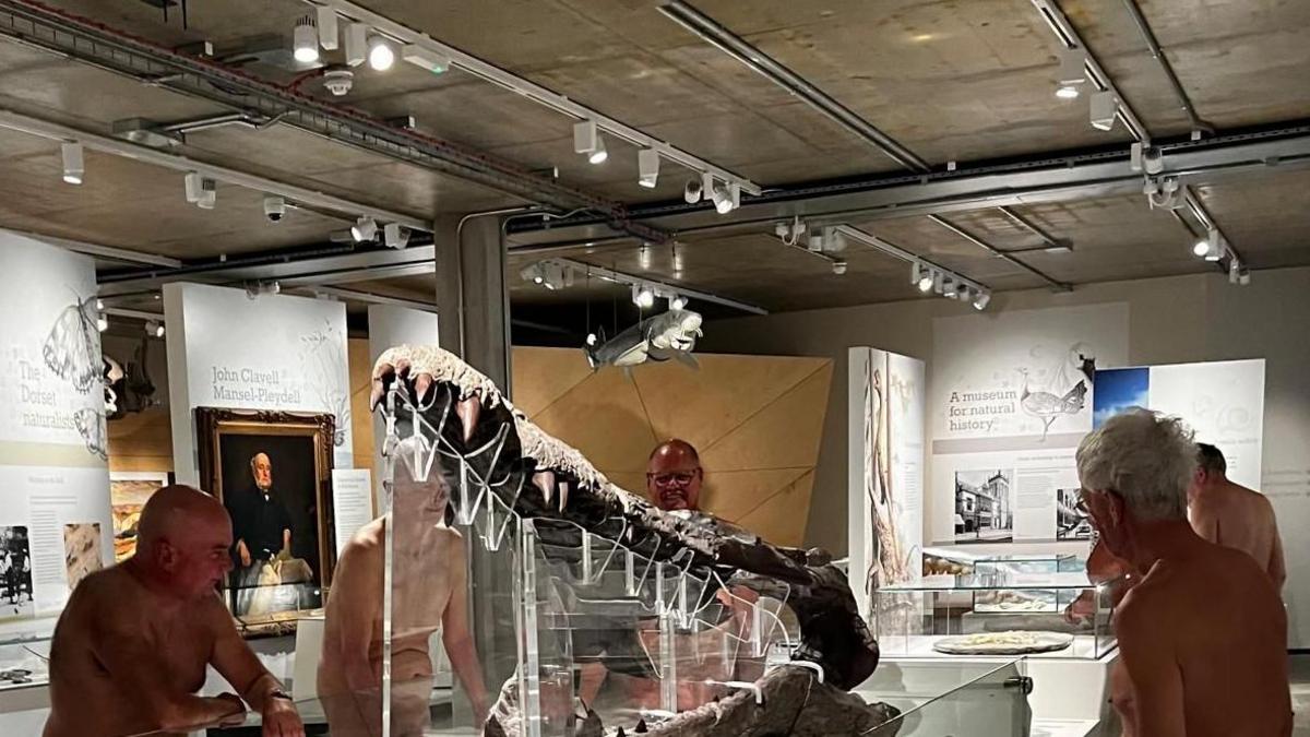 A group of naked men surrounding a large crocodile-like fossil exhibit in a museum gallery 