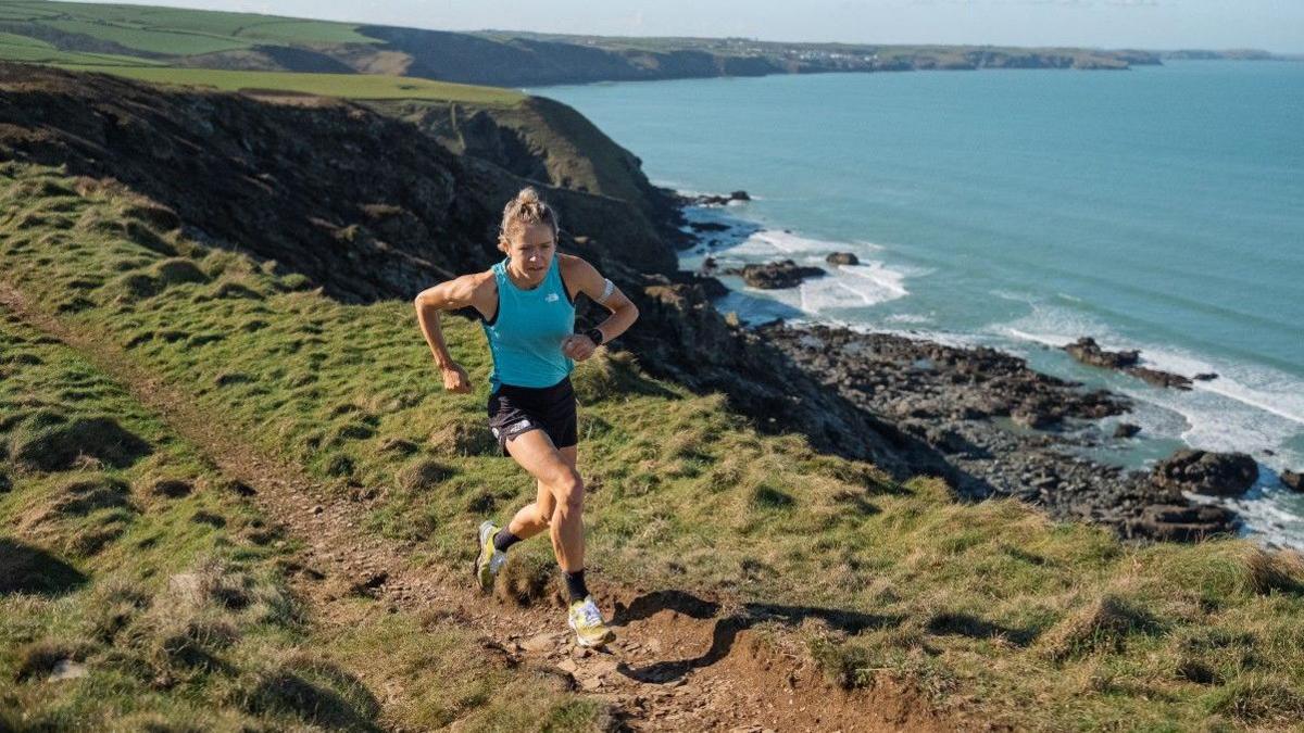 Elsey Davis runs along a clifftop wearing black shorts and a blue vest top. In the background you can see the sea and coastline on a sunny day. 
