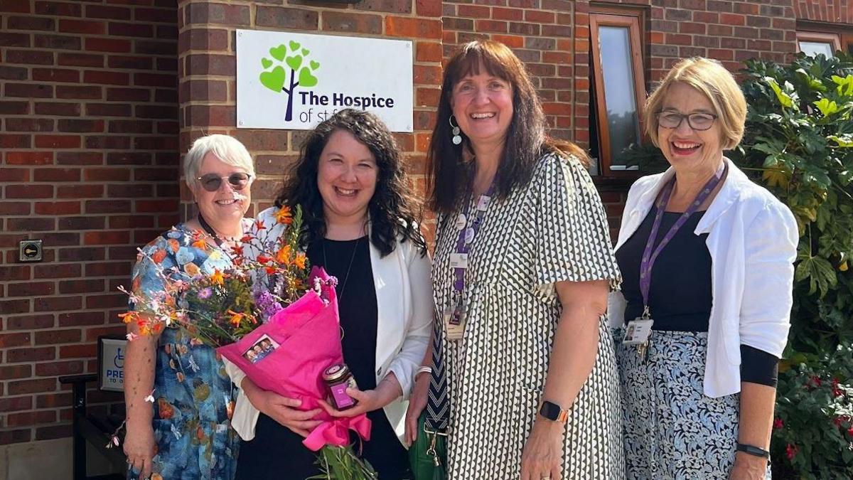 Three ladies in summer outfits pose with MP Victoria Collins outside the entrance of the hospice. Ms Collins is holding a bunch of flowers and a jar of preserve. 