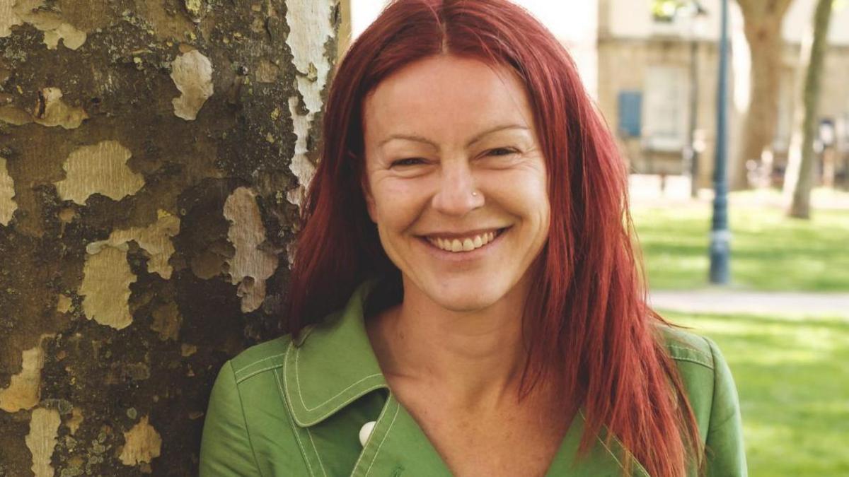 Christine Townsend with red hair, leaning against a tree and smiling at the camera