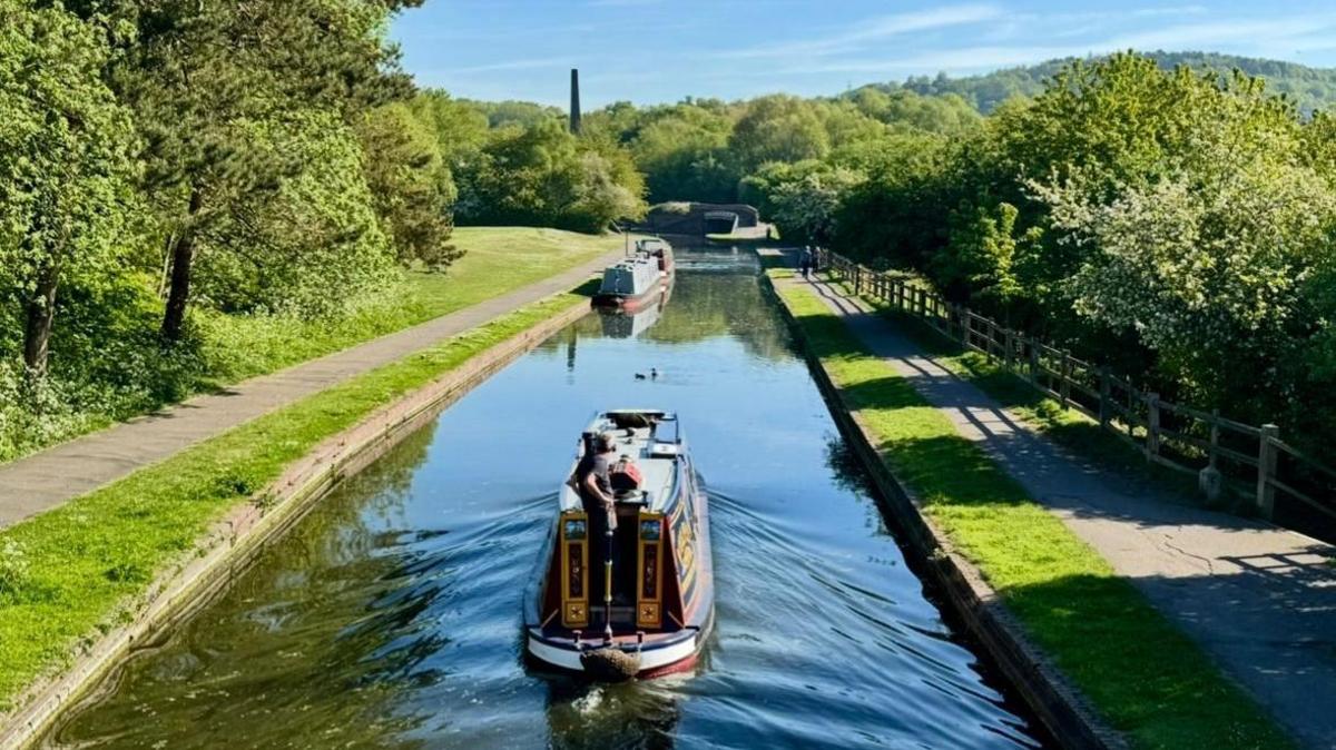 A narrowboat moves away from us along a canal, leaving a water wake behind it. a person can just about be seen at the back holding the rudder. The boat has white, red and yellow trim. Either side of the canal are green, grassy banks with trees beyond towpaths.