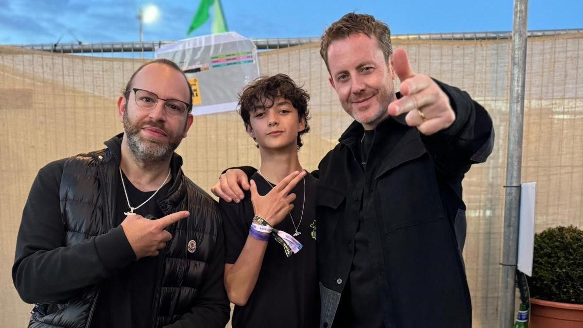 A young boy in a black T-shirt posting for a photo with two men, both wearing black jackets. They're standing in front of a temporary festival fence and the sky is blue.