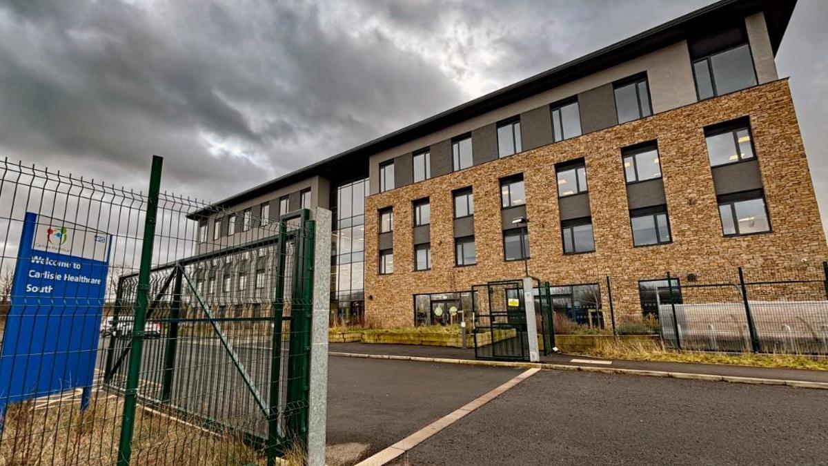 Side view of a large four storey building - a health centre - with rows of windows and a glassed-in area in the centre. There is wire fencing around it, with a large open gate for the access road. A sign to the left reads: Welcome to Carlisle Healthcare South