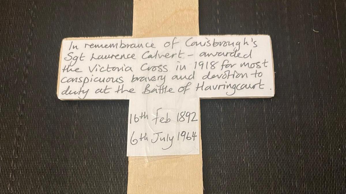 A close-up of the cross planted - a piece of paper is taped to it. A handwritten message on the paper reads: "In remembrance of Conisbrough's Sgt Laurence Calvert - awarded the Victoria Cross in 1918 for most conspicuous bravery and devotion to duty at the Battle of Havrincourt. 16th Feb 1892. 6th July 1964."