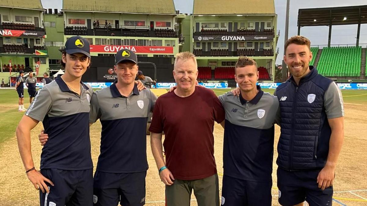 Alan Davis is stood in the middle of four Hampshire cricket players. They are stood in front of a wicket and a large grandstand.