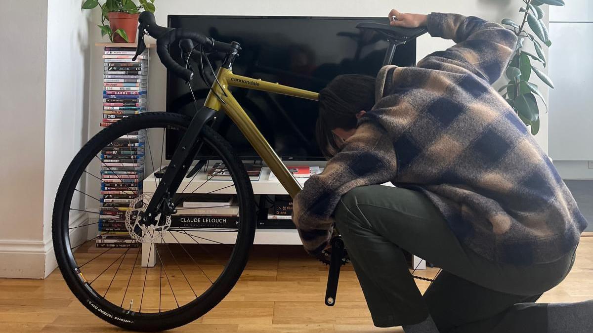 Alasdair working on his bike in his home. He is knelt down using a tool by the back wheel. 