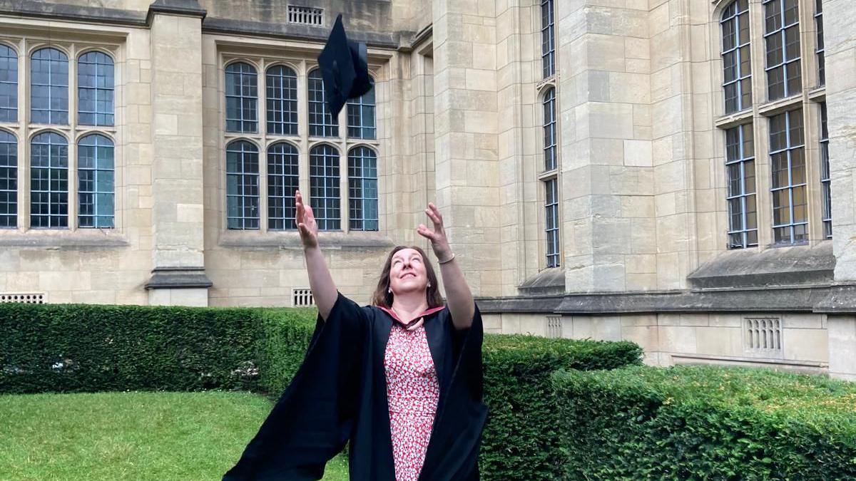 Caroline throws her graduation hat in the air, wearing the graduation robes and a pink dress, standing on the lawn in front of a university building 