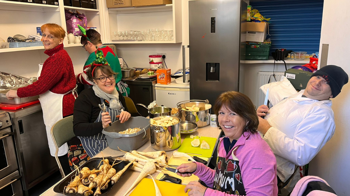 Five charity volunteers, some wearing festive hats, are either standing in a kitchen or sitting at a table preparing parsnips for cooking in large silver saucepans