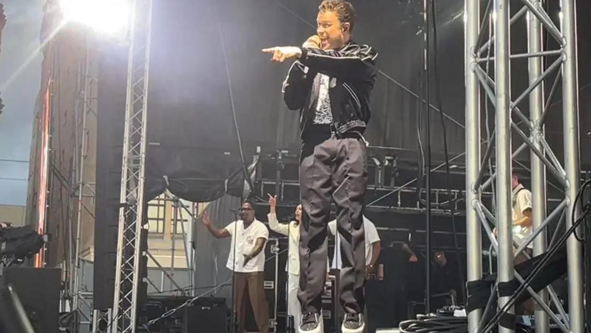 Olly Murs is singing and jumping in the air on stage, with his band in the background. He is smiling and pointing at the crowd.