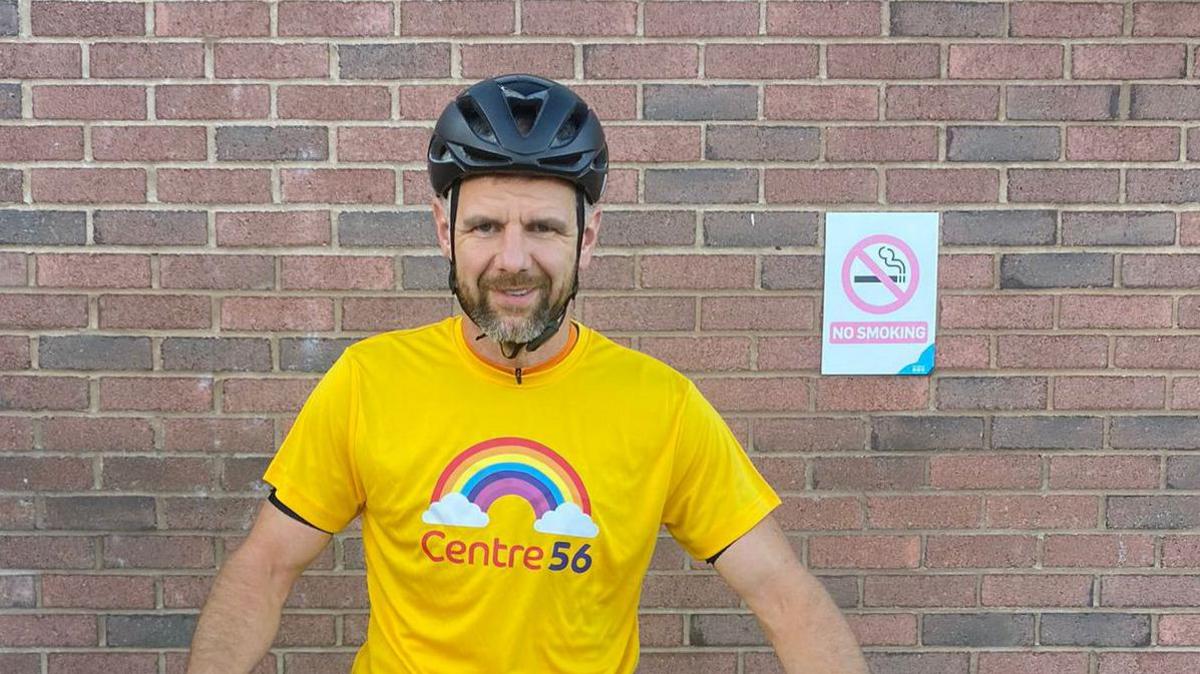 Mickey Burns, who has a brown beard with grey flecks, is wearing a black cycling helmet and yellow t-shirt with the Centre 56 charity logo of a rainbow and two clouds on it. He is standing in front of a brick wall, smiling for the camera.