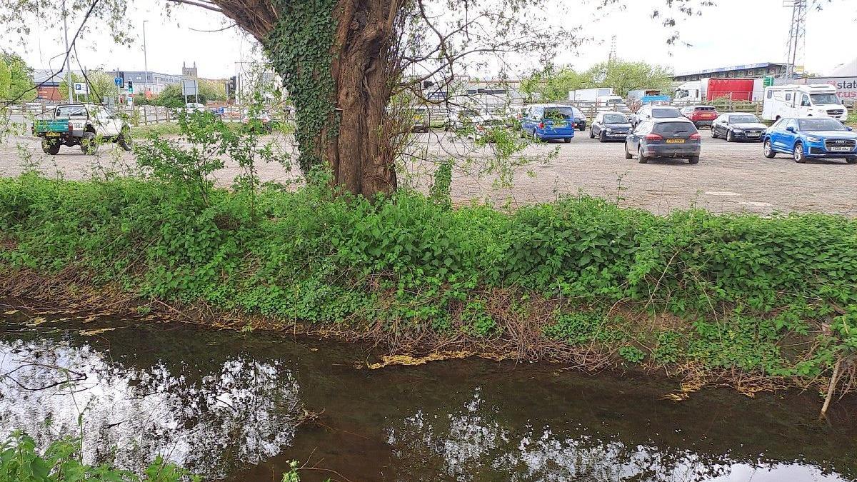 A shallow brook runs adjacent to a small car park with Hereford FC's stadium in the background. 