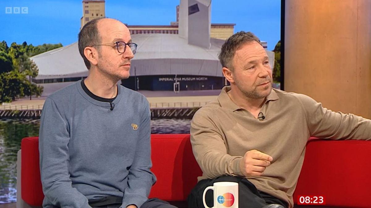 Jack Thorne (left) and Stephen Graham sit on a red sofa in the BBC studio. Jack wears a blue jumper, he has short hair and wears black-rimmed glasses. Stephen wears a light brown collared jumper.