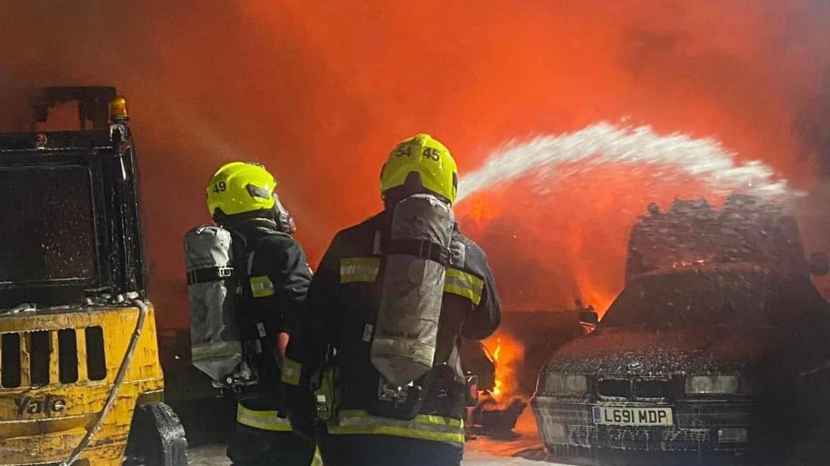 Firefighters spray water on car with fire in background