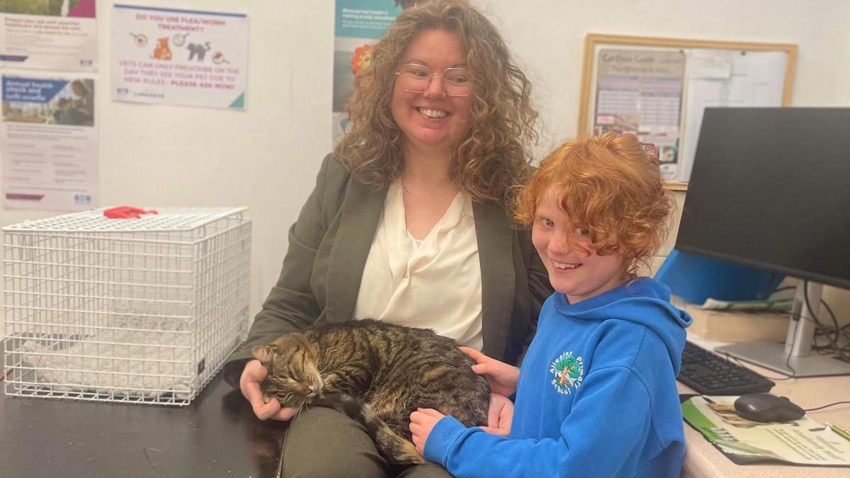 A woman sits in a vetinary practice with a tabby cat on her lap. A child stands next to her stroking the cat.