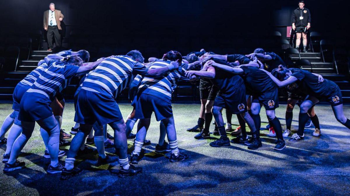 A rugby scrum is shrouded in blue light in the centre of the stage that has been turned into Sandy Park the home ground of the Exeter Chiefs.