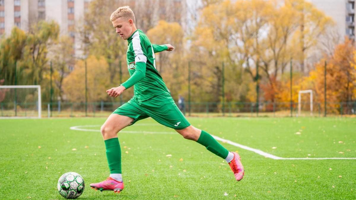 A young teenager with blonde hair, wearing a green football kit, kicks a football on a football pitch 