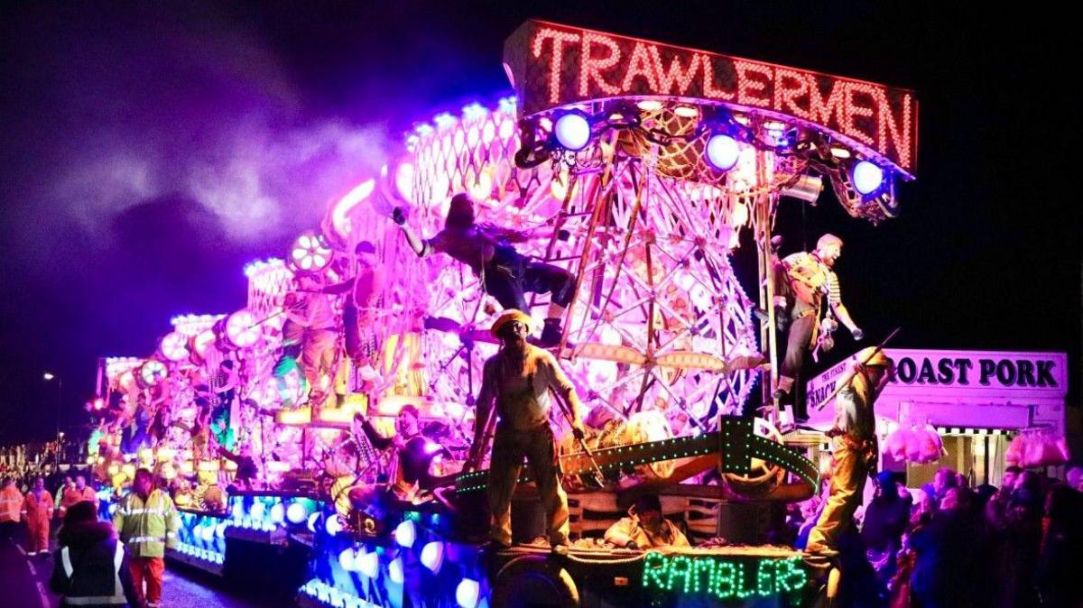 An illuminated carnival cart with purple, blue and red lights. There are people standing on it and a crowd gathering around it.