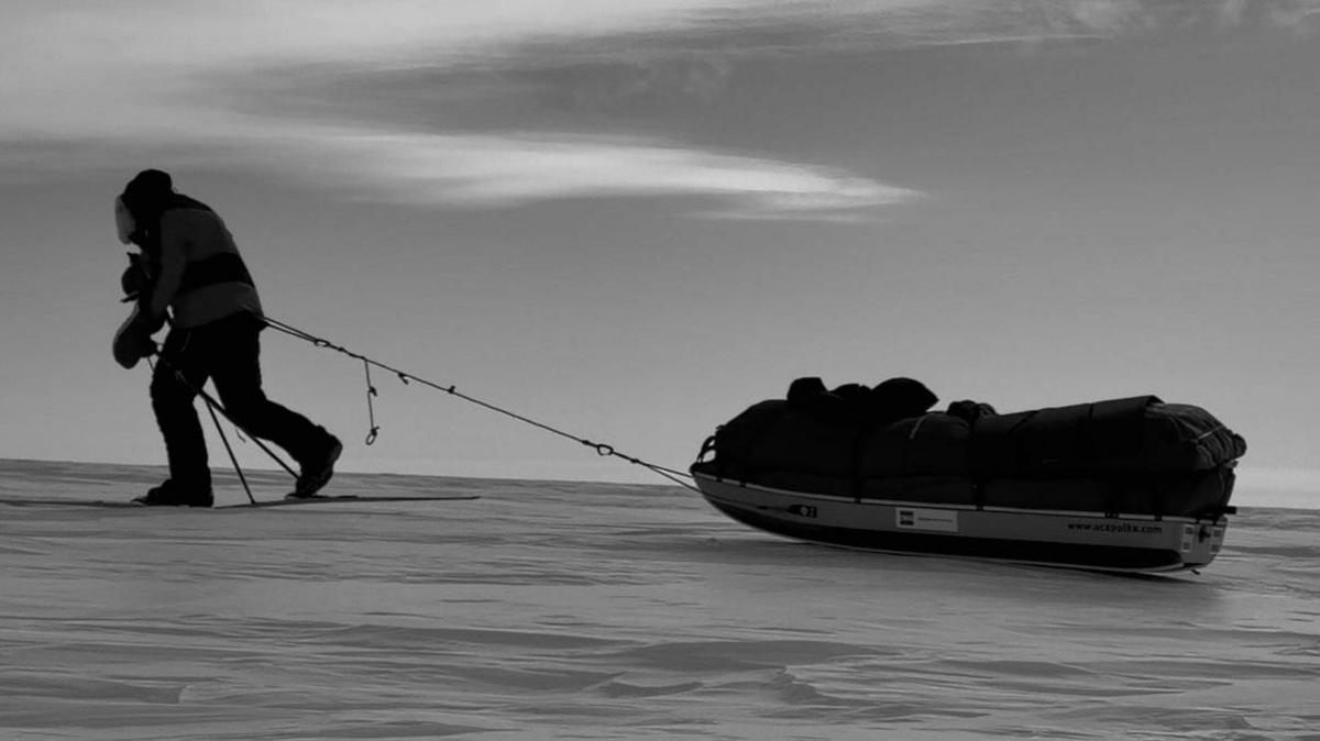 Black and white side profile shot of Ian in his furn lined hooded coat and trousers pulling a sled with baggage on it. He is on a flat section of ice.