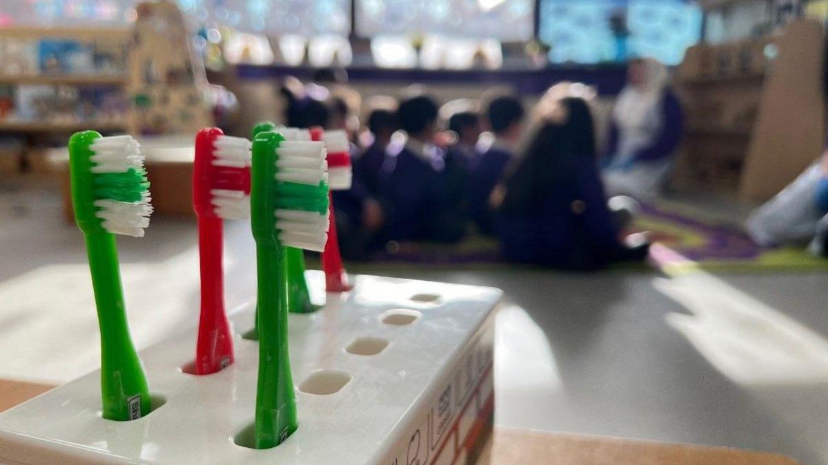 In the foreground there is a toothbrush holder. Three green and two red toothbrushes are visible. Children, who are blurred, are sitting in the background.