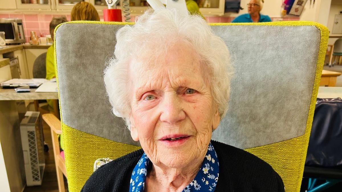 Millie Donnelly, a 99-year-old care home resident sitting in a yellow chair, with other people in the background