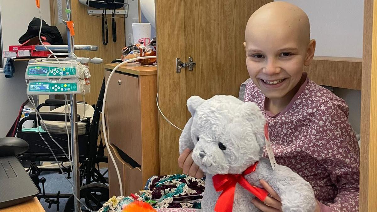 Poppi in a hospital bed cuddling a large teddy bear. She is wearing red pyjamas with a white pattern and is smiling. She has lost her hair because of her treatment. There is a medical machine and a chest of drawers next to the bed.